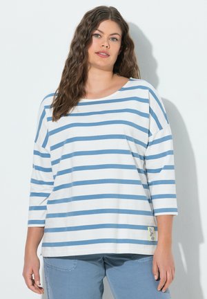 Frau mit langen, welligen Haaren trägt ein weiß-blau gestreiftes Langarmshirt und blaue Hosen und steht vor weißem Hintergrund.