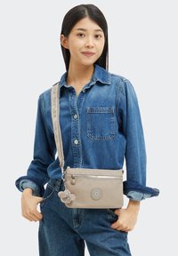 Junge Frau, die ein Jeanshemd und Jeans trägt, während sie eine beige Umhängetasche mit einem plüschigen Schlüsselanhänger und einem gebrandeten Riemen trägt.