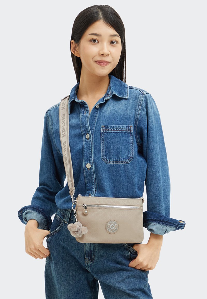 Junge Frau, die ein Jeanshemd und Jeans trägt, während sie eine beige Umhängetasche mit einem plüschigen Schlüsselanhänger und einem gebrandeten Riemen trägt.