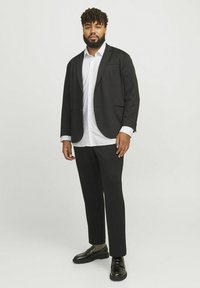 Schwarze Jacke mit einer Tasche, weißes Hemd, schwarze Hosen und schwarze Slipper. Glatter Stoff mit taillierter Passform.