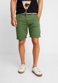 Shorts en coton verts avec des ourlets retroussés, des poches latérales et un bouton marron comme accent. Associés à des baskets blanches et un t-shirt graphique noir.