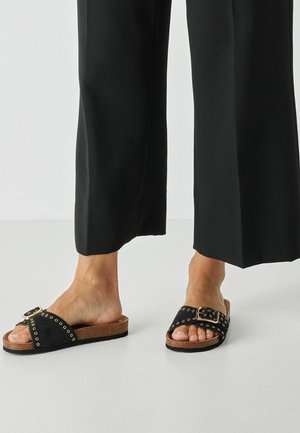 Sandales à slide noires avec une large bride ornée d'une grande boucle et de clous métalliques, posées sur une semelle en liège pour plus de confort.