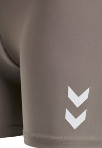 Close-up van taupe sportshorts met twee witte naar beneden gerichte chevron-logo's dicht bij de zoom op gladde stof.