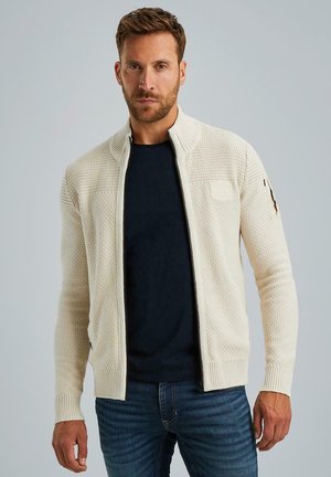 Cream knit sweater with a zip, textured pattern, high collar, ribbed cuffs, and a patch on the sleeve, worn over a dark t-shirt and jeans.