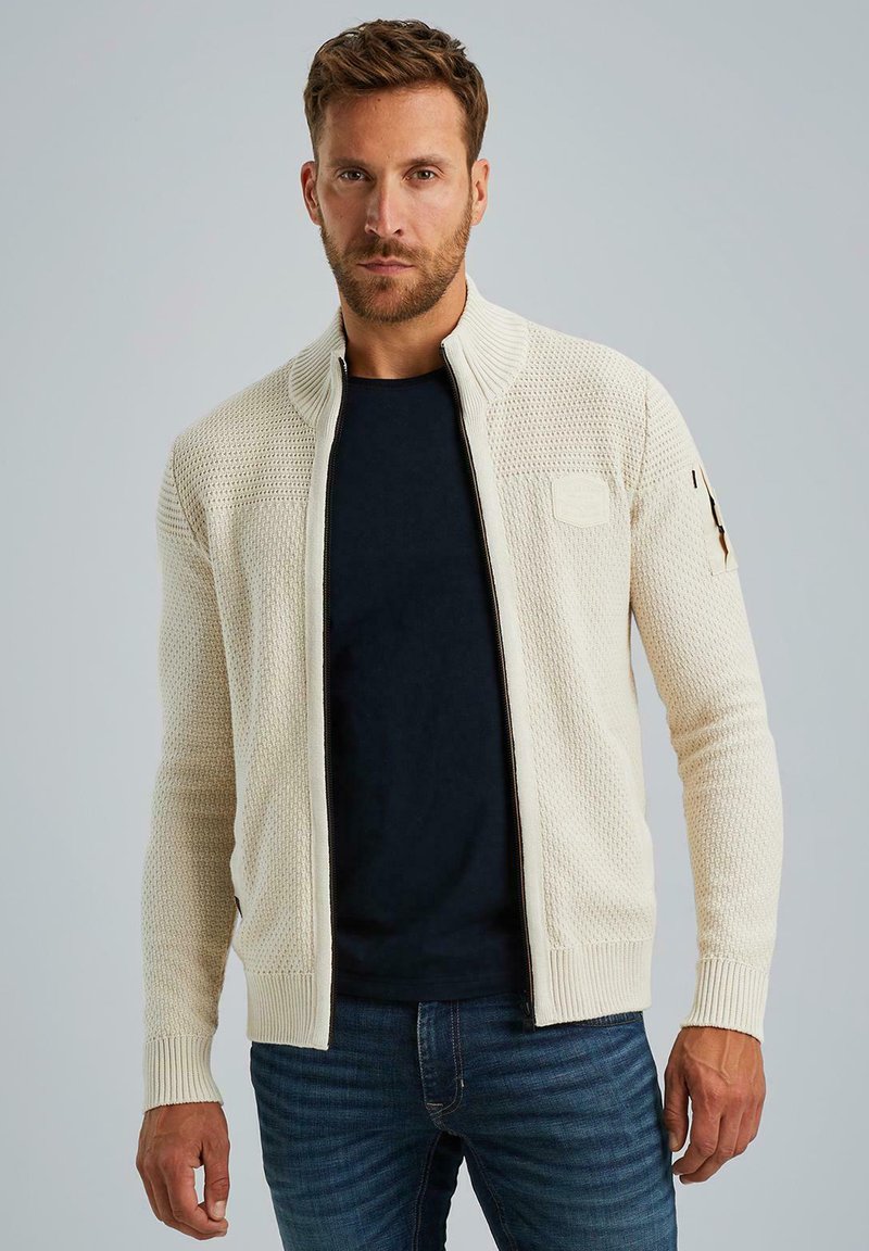 Cremefarbener Strickpullover mit Reißverschluss, strukturiertem Muster, hohem Kragen, gerippten Bündchen und einem Patch am Ärmel, getragen über einem dunklen T-Shirt und Jeans.