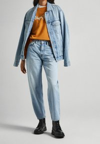 Ljusblå jeansjacka med två framfickor, bärs över en orange sweatshirt med vit text. Ljusblå jeansbyxor med avslappnad passform och svarta kängor.