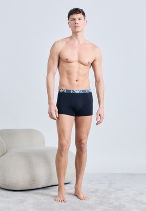 Jeune homme debout pieds nus en boxer noir avec ceinture de marque, expression neutre, à côté d'une chaise moderne gris clair sur un tapis.