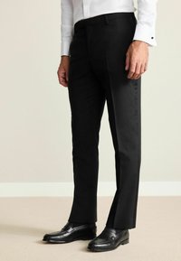 Schwarze angepasste Hosen mit glatter Oberfläche, geradem Schnitt und seitlichen Taschen, kombiniert mit schwarzen Lederloafers. Weißes Hemd ist auf Taillenhöhe sichtbar.