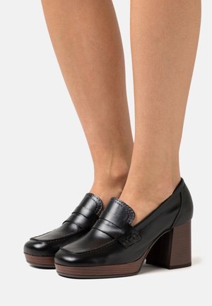 Mocassins en cuir noir à talon bloc avec un bout arrondi, des coutures décoratives et une texture lisse. Talon à texture bois qui crée un contraste.