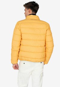 Chaqueta de plumas amarilla con costuras horizontales, cuello alto y logotipo en la parte trasera. Cuenta con mangas largas y una silueta ajustada.