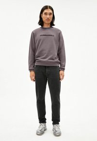 Grijze trui met zwarte "ARMEDANGELS" tekst, geribbelde halslijn en manchetten. Gecombineerd met zwarte spijkerbroek en grijze sneakers. Gladde stoftextuur.