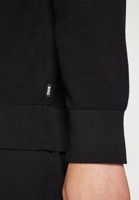 Gros plan sur la manche et le poignet d'un pull en maille noir avec une petite étiquette de la marque BOSS, reposant au-dessus d'un pantalon noir et d'un poignet visible.