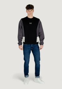 Maglione nero e grigio con vestibilità oversized, dotato di scollo tondo e logo. Abbinato a jeans blu e sneaker bianche.