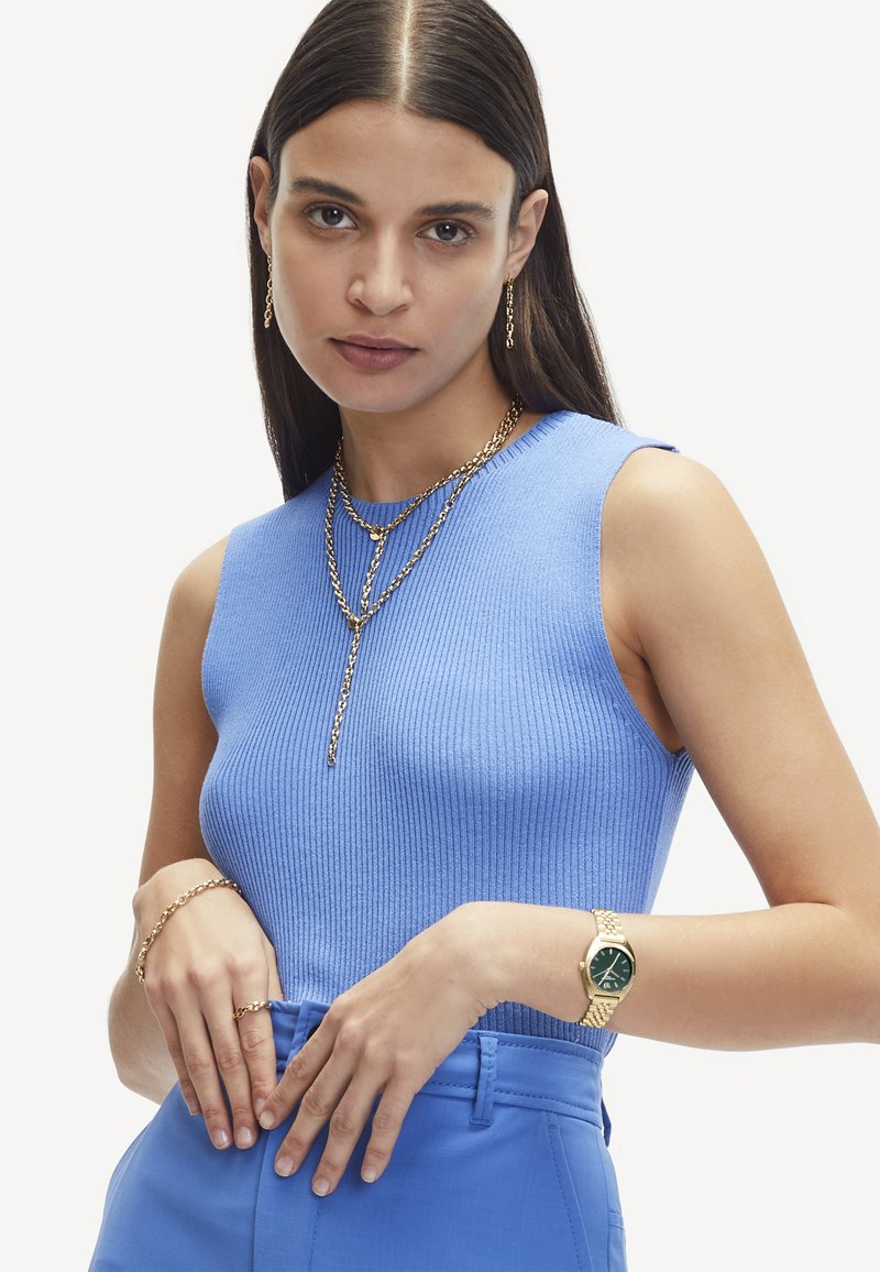 Lichtblauw geribbeld mouwloos topje met ronde hals, gecombineerd met blauwe broek. Gouden gelaagde ketting en horloge met groene wijzerplaat.