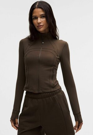 Barna, cipzáros sportdzseki hosszú ujjakkal, testhezálló szabással és texturált anyagból; oldalán zsebekkel és kontrasztos varrásokkal.