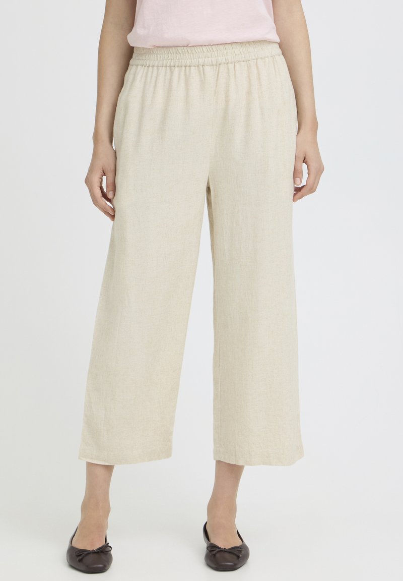 Femme portant un pantalon beige à jambes larges et courtes avec une ceinture élastique, associé à des ballerines noires, debout devant un fond uni.