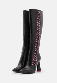 Botas de cuero negras hasta la rodilla con cordones laterales rosas, punteras afiladas y tacones acampanados con suelas rosas, mostradas de pie juntas sobre un fondo blanco.