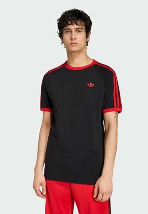 Mladý muž v černém tričku Adidas s červenými pruhy a logem, v kombinaci s červenými kalhotami, stojící před jednoduchým světlým pozadím.