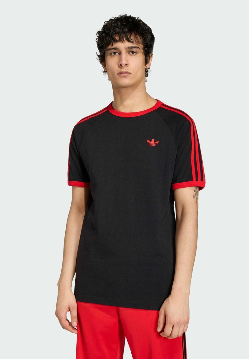 Jeune homme portant un t-shirt Adidas noir avec des rayures et un logo rouges, associé à un pantalon rouge, debout devant un fond clair uni.