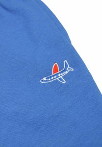 Chemise en coton bleu avec un petit design d'avion brodé en blanc, rouge et bleu sur la manche. Texture lisse avec une coupe décontractée.