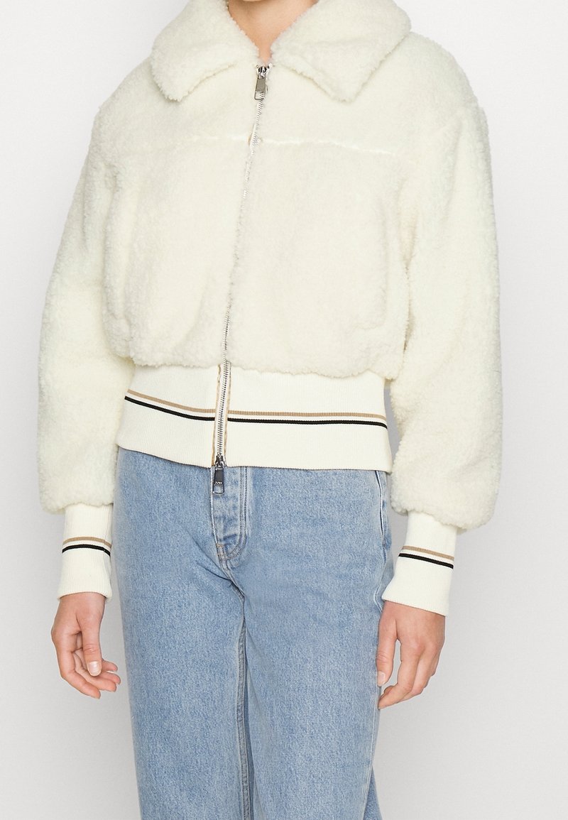 Person som bär en kort, vit sherpa zip-up jacka med ribbade muddar och fåll, tillsammans med ljusblå högmidjade jeans.