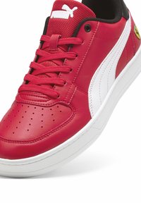 Adidași din piele roșie cu accente albe, guler negru și vârf perforat. Prezintă o talpă texturată și logo-uri proeminente Puma și Ferrari.