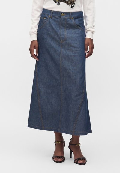 Femme portant une longue jupe en jean foncé avec des poches devant, un pull blanc et des sandales noires à talons hauts avec des brides à la cheville.