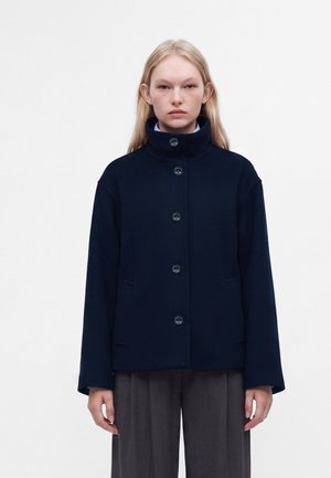 Jeune femme aux longs cheveux blonds portant une veste bleu marine boutonnée à col montant et un pantalon gris ample, debout devant un fond uni.