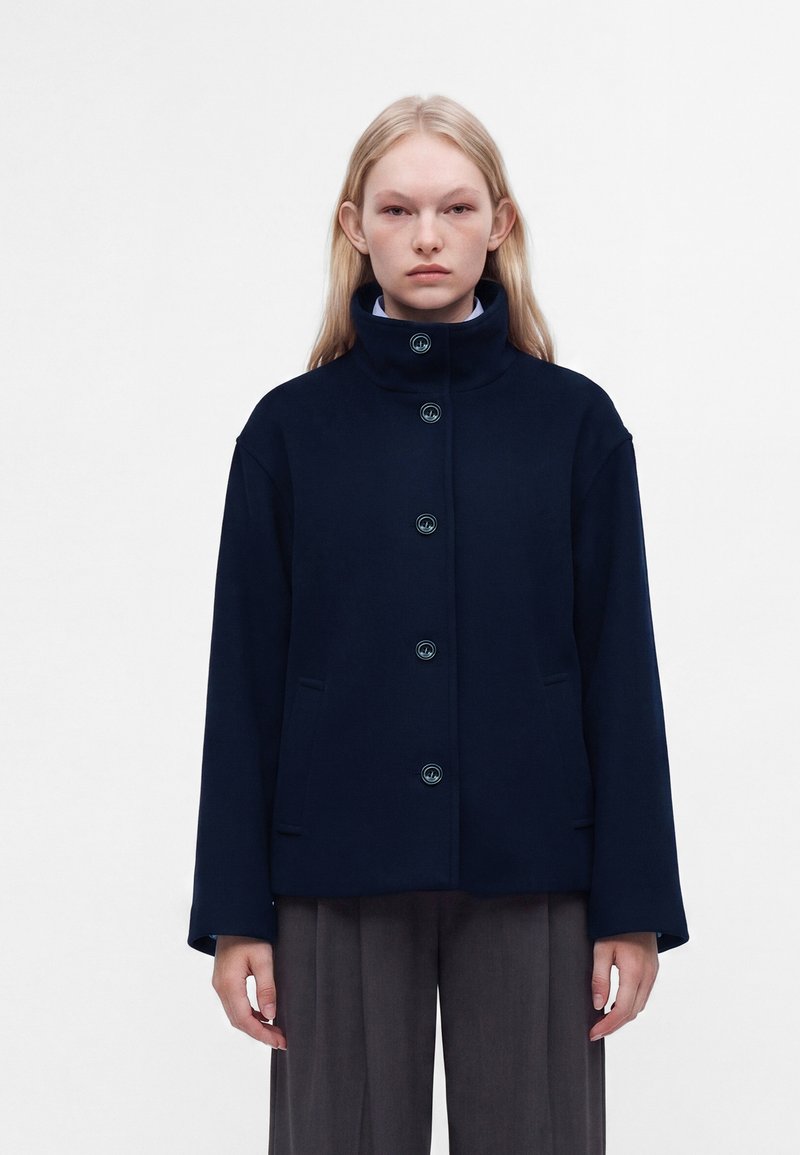 Jeune femme aux longs cheveux blonds portant une veste bleu marine boutonnée à col montant et un pantalon gris ample, debout devant un fond uni.