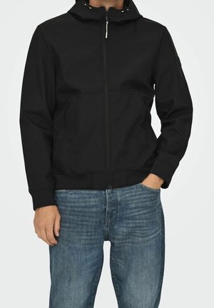 Uomo con giacca nera con cappuccio e zip e jeans blu, in piedi con una mano in tasca su sfondo uniforme.