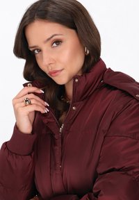 Giubbotto imbottito bordeaux con colletto alto, chiusura con zip e accessori in tonalità oro. Presenta polsini a coste e una finitura liscia e lucida.