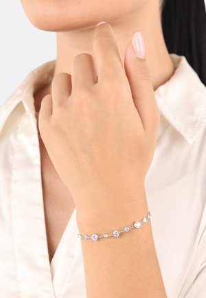 Delicata bracciale d'argento con gemme rotonde e ovali equidistanti indossato sul polso, mano vicino al mento, colletto di camicia bianca visibile.