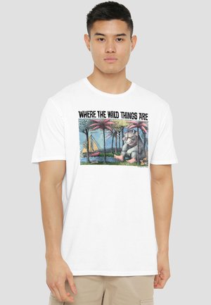Young man wearing white t-shirt with "Where the Wild Things Are" text and illustration of a creature and sailboat among palm trees.