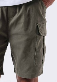 Bermudas cargo verde oliva hechas de tela texturizada, con bolsillos laterales y un corte relajado. Costuras limpias y detalles en el dobladillo visibles.