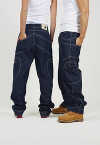 Temno modri denim cargo hlače z več žepi, kontrastnim šivanjem in ohlapnim krojem, v kombinaciji s svetlo rjavimi čevlji. Ravno krojen stil.