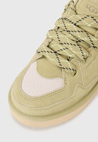 Lichtgroene suède sneaker met een getextureerde roze mesh accent, voorzien van gestreepte veters en een rubberen zool. Close-up van het teen vlak.
