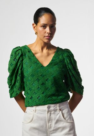 Vrouw met donker haar naar achteren gebonden, draagt gouden oorbellen, een groene gestructureerde blouse met pofmouwen en hoge witte broek tegen een witte achtergrond.