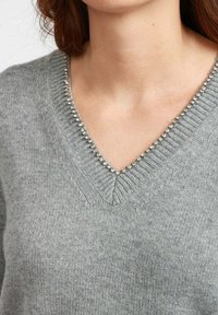 Maglione grigio a V lavorato a maglia con piccole strass trasparenti che decorano il colletto, indossato da una persona con lunghi capelli castani.