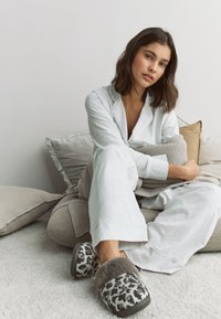 Ensemble de pyjama gris clair à manches longues ; chaussons en peluche à imprimé léopard avec des détails en fausse fourrure grise ; assis sur une surface douce et texturée avec des coussins.
