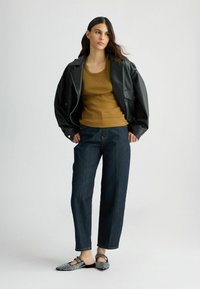 Giacca di pelle nera sopra una canotta a coste senape, abbinata a jeans ampi in denim scuro e ballerine stampate con decorazioni.