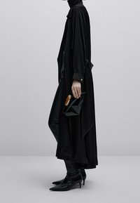 Długi czarny płaszcz o eleganckim fasonie, zestawiony z dopasowanymi czarnymi spodniami i szpiczastymi czarnymi szpilkami. Trzyma geometryczną czarną torebkę z złotym akcentem.