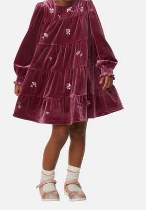 Niño vestido con un vestido de terciopelo de manga larga y volantes, con pequeños bordados florales, calcetines blancos texturizados y zapatos rosas con detalles en colores del arcoíris.