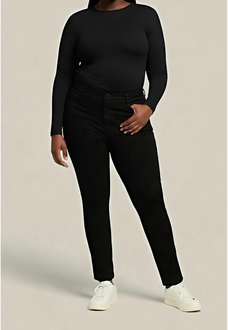 Personne portant un haut noir ajusté à manches longues, un pantalon noir taille haute et des baskets blanches, debout avec une main dans la poche.