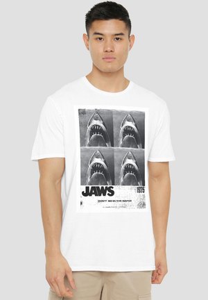 Young man wearing white T-shirt with black-and-white four-panel shark graphic and text "JAWS 1975 DON'T GO IN THE WATER."