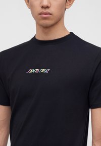Άνδρας με μαύρο κοντομάνικο T-shirt και πολύχρωμο κείμενο "Santa Cruz" τυπωμένο στο στήθος