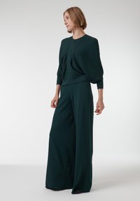 Donkergroene, gestructureerde top met geplooid ontwerp, gecombineerd met een wijde broek in dezelfde stof. De outfit heeft een relaxte pasvorm en lange mouwen.