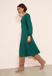 Rochie verde texturată cu decolteu în V și mâneci bufante, asortată cu cizme de piele suede, bej, până la genunchi. Material neted, design de lungime medie.
