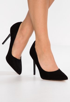 Fødder iført sorte ruskindspointe hæle med slanke stiletto hæle på en hvid gulvbaggrund.