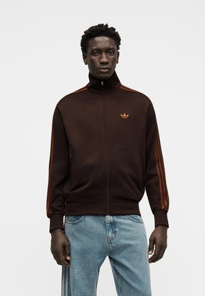 Uomo con giacca sportiva Adidas marrone scuro con zip, strisce arancioni e logo, abbinata a jeans azzurri, in piedi davanti a uno sfondo bianco.