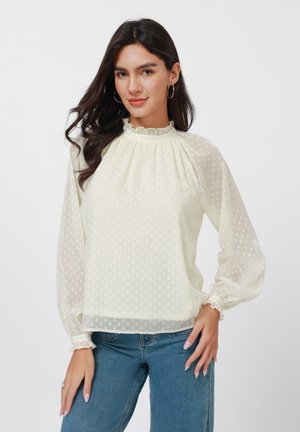 Hellgelbe Bluse mit Punkten, gerüschtem Ausschnitt und Puffärmel. Aus transparenter Stoff gefertigt, kombiniert mit Jeans.